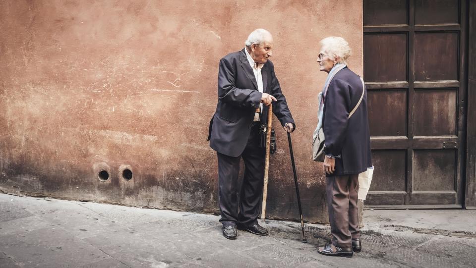 Ein älterer Mann mit Gehstock und eine ältere Frau unterhalten sich vor einer Hauswand